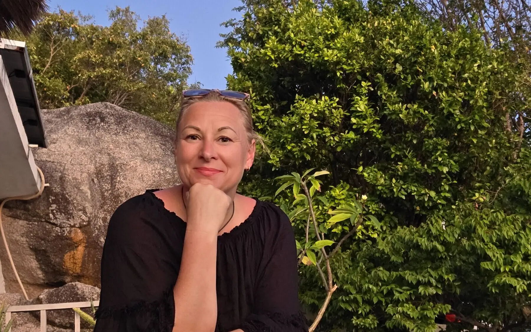Heike Berndt sitzt draußen auf einer Steinmauer unter Bäumen